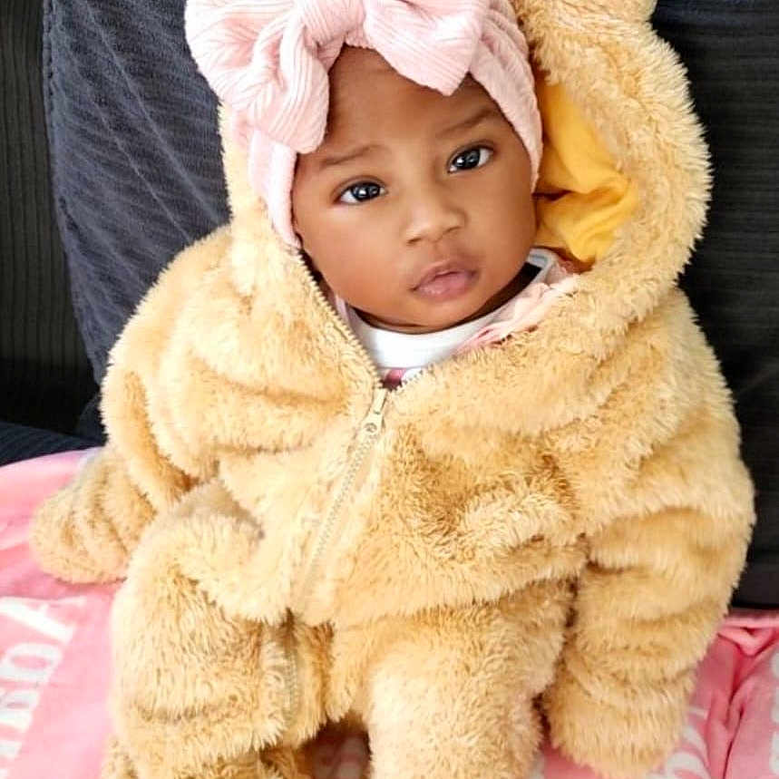Adanna joined the competition — help win amazing prizes! adorable, baby, blanket, child, clothing, cozy, cushion, cute, expression, face, fluffy_clothing, headband, indoor, infant, pink_bow, portrait, seated, soft_texture, teddy_bear_onesie, warm