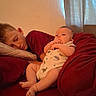 baby, child, sleeping, sibling, bed, blanket, pillow, curtain, indoor, onesie, dinosaur_pattern, red_blanket, resting, infant, person, family, cozy, relaxing, natural_light, casual