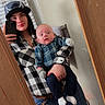 River is registered to the contest to win money with this photo: woman, baby, mirror, selfie, cap, plaid_shirt, blue_pants, bathroom, towel, door, wood, floor, phone, reflection, hand, face, smile, holding, jeans, gloves