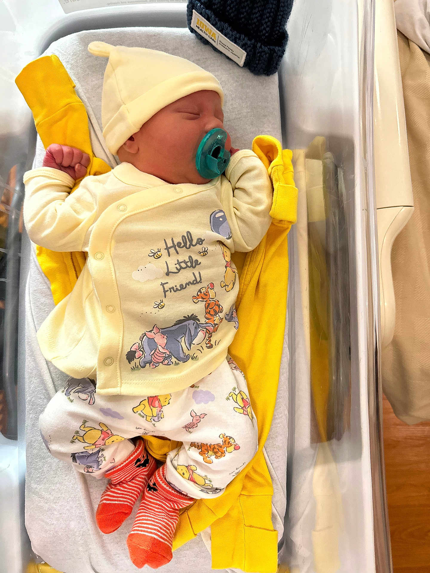 River is registered to the contest to win money with this photo: newborn, baby, infant, sleeping, pacifier, hospital_bassinet, yellow_clothing, hat, socks, winnie_the_pooh, cartoon_characters, child, cute, cozy, bed, blanket, person, indoors, resting, soft