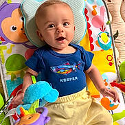 Zeus is registered to the contest to win money with this photo: baby, baby_gear, blue_clothing, child, colorful, elephant_toy, fish_shirt, happy, indoor, infant, looking_at_camera, lying_down, musical_toys, play_mat, playroom, portrait, smiling, stuffed_animal, toy, yellow_pants