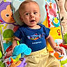 baby, baby_gear, blue_clothing, child, colorful, elephant_toy, fish_shirt, happy, indoor, infant, looking_at_camera, lying_down, musical_toys, play_mat, playroom, portrait, smiling, stuffed_animal, toy, yellow_pants