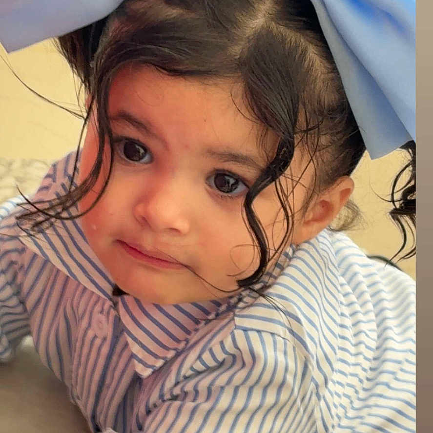 Donaima participe au concours pour gagner de l'argent avec cette photo : big_eyes, blue_bow, button, child, clothing, curly_hair, cute, expression, face, hair, indoor, light, mobile_screen, person, portrait, social_media, story, striped_shirt, toddler, young_child