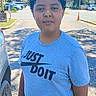 boy, buildings, car, casual_clothing, child, curly_hair, daytime, outdoor, parking_lot, person, road, shorts, sidewalk, sky, standing, sunlight, trees, tshirt, urban, vehicle
