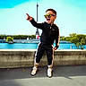 child, boy, sunglasses, tracksuit, sneakers, pointing, outdoor, eiffel_tower, landmark, river, concrete_wall, trees, blue_sky, happy, smiling, fashion, urban, daylight, person, portrait