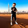 Yanis is registered to the contest to win money with this photo: child, boy, tracksuit, peace_sign, desert, sand, sky, blue_sky, sunlight, shoes, urban_skyline, skyscraper, city, outdoor, portrait, smile, standing, fashion, casual_wear, young