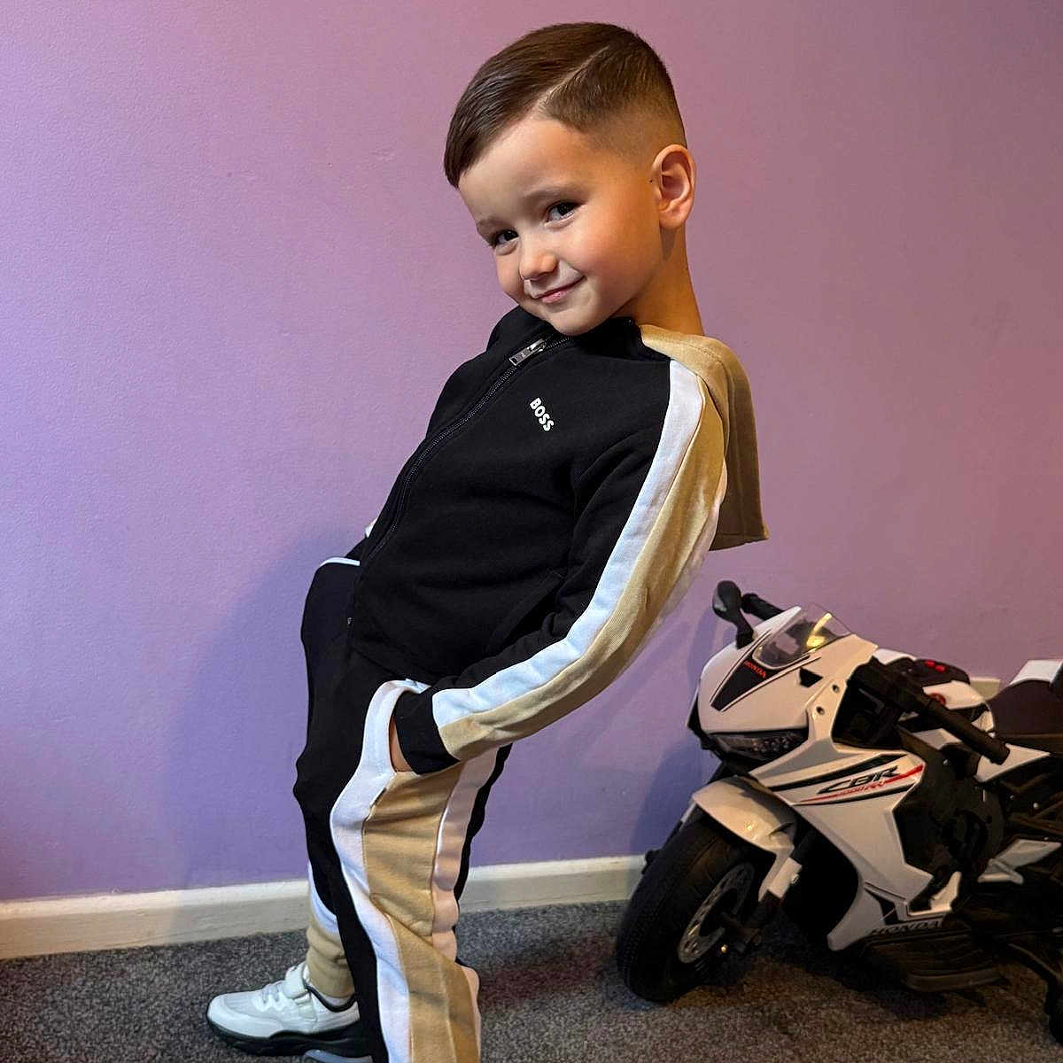 Yanis is registered to the contest to win money with this photo: child, boy, pose, tracksuit, black, beige, sneakers, indoor, purple_wall, toy_motorcycle, carpet, smile, haircut, playful, confident, floor, young, person, casual, fashion