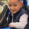 child, boy, smile, amusement_ride, blue_car, indoor, portrait, haircut, vest, striped_shirt, playful, face, person, entertainment, fun, leisure, technology_overlay, app_interface, happy, kid