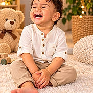 Arshin Khamisi joined the competition — help win amazing prizes! child, toddler, smiling, laughing, portrait, curly_hair, sitting, cross_legged, indoor, cozy, warm_lighting, white_shirt, beige_pants, barefoot, fluffy_rug, teddy_bear, houseplant, string_lights, nursery, happy