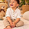 child, toddler, smiling, laughing, portrait, curly_hair, sitting, cross_legged, indoor, cozy, warm_lighting, white_shirt, beige_pants, barefoot, fluffy_rug, teddy_bear, houseplant, string_lights, nursery, happy