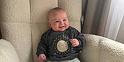 Vinnie is registered to the contest to win money with this photo: armchair, baby, child, cozy, curtains, cute, face, furniture, happy, indoor, infant, light, pants, plush_furniture, portrait, seated, smile, socks, soft_light, sweatshirt