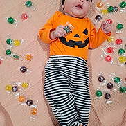 Arabella is registered to the contest to win money with this photo: baby, black_and_white, black_headband, blanket, bow, candy, child, colorful, costume, cute, footwear, halloween, indoors, infant, lying_down, orange_shirt, pumpkin_face, smile, striped_pants, wrapped_candy