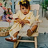 toddler, child, yellow_coat, floral_dress, wooden_chair, keys, curly_hair, hair_bows, stuffed_bunny, basket, flowers, indoor, portrait, cute, curious, floor_rug, decor, baby_sandals, seated, expression