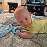 baby, infant, child, tummy_time, smiling, portrait, yellow_clothing, toy, turquoise_toy, bed, bedspread, indoor, living_room, television, furniture, blanket, playtime, face, happy, photography