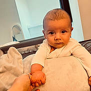 Maverick is registered to the contest to win money with this photo: baby, infant, child, hand_holding, cushion, white_clothing, indoor, soft_light, face, portrait, looking_at_camera, furniture, blanket, adult_hand, skin, expression, cozy, home, interaction, closeup