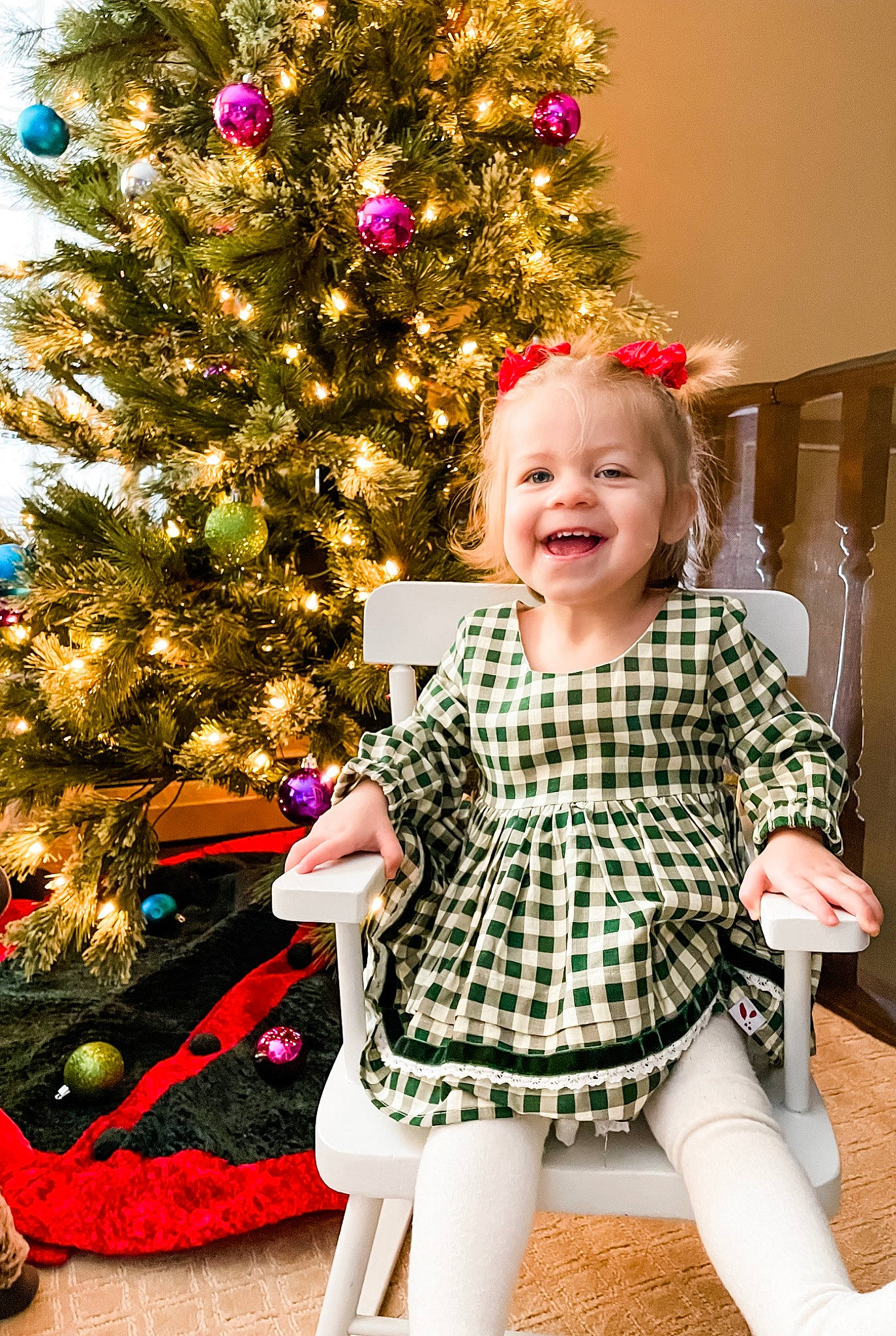 Adelynn joined the competition — help win amazing prizes! baby_toddler_clothing, christmas_ornament, christmas_tree, dress, facial_expression, happy, head, holiday_ornament, human_body, joy, ornament, person, photograph, pink, plaid, plant, sleeve, smile, standing, tartan