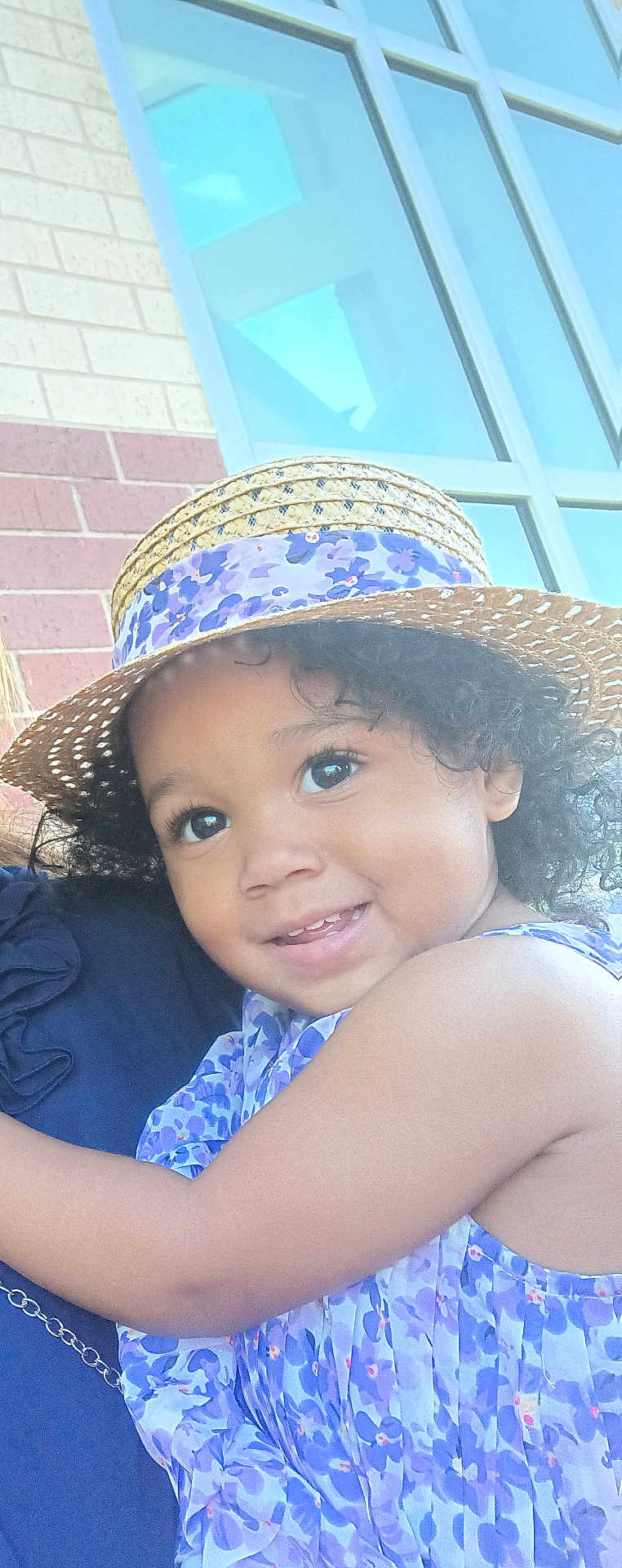 Khaycee joined the competition — help win amazing prizes! toddler, child, smiling, hat, straw_hat, purple_dress, floral_pattern, curly_hair, outdoor, window, brick_wall, face, happy, portrait, person, sunlight, cute, closeup, skin, arm