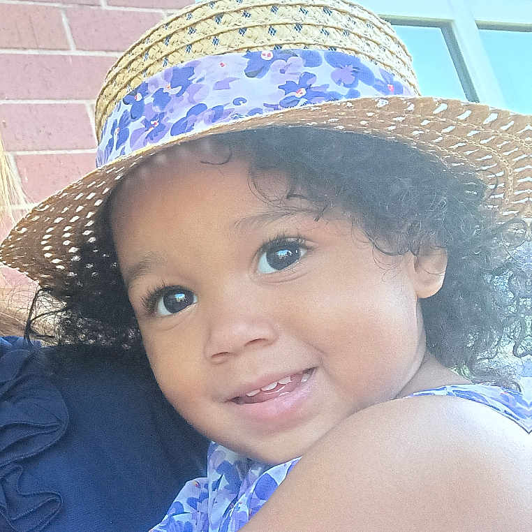 Khaycee joined the competition — help win amazing prizes! brick_wall, casual, child, curly_hair, cute, eyes, face, fashion, happy, hat, hugging, outdoor, person, portrait, purple_dress, smiling, straw_hat, sunlight, toddler, window