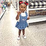 blue_dress, child, confident, expression, floor, girl, home_goods, indoor, person, polka_dot, pose, red_bows, retail, shelves, shopping, standing, store_aisle, toddler, white_shoes, young_child