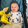baby, infant, stroller, blanket, towel, plush_toy, yellow, dinosaur_pattern, onesie, smile, face, child, soft_texture, cozy, indoor, person, cute, clothing, comfort, resting