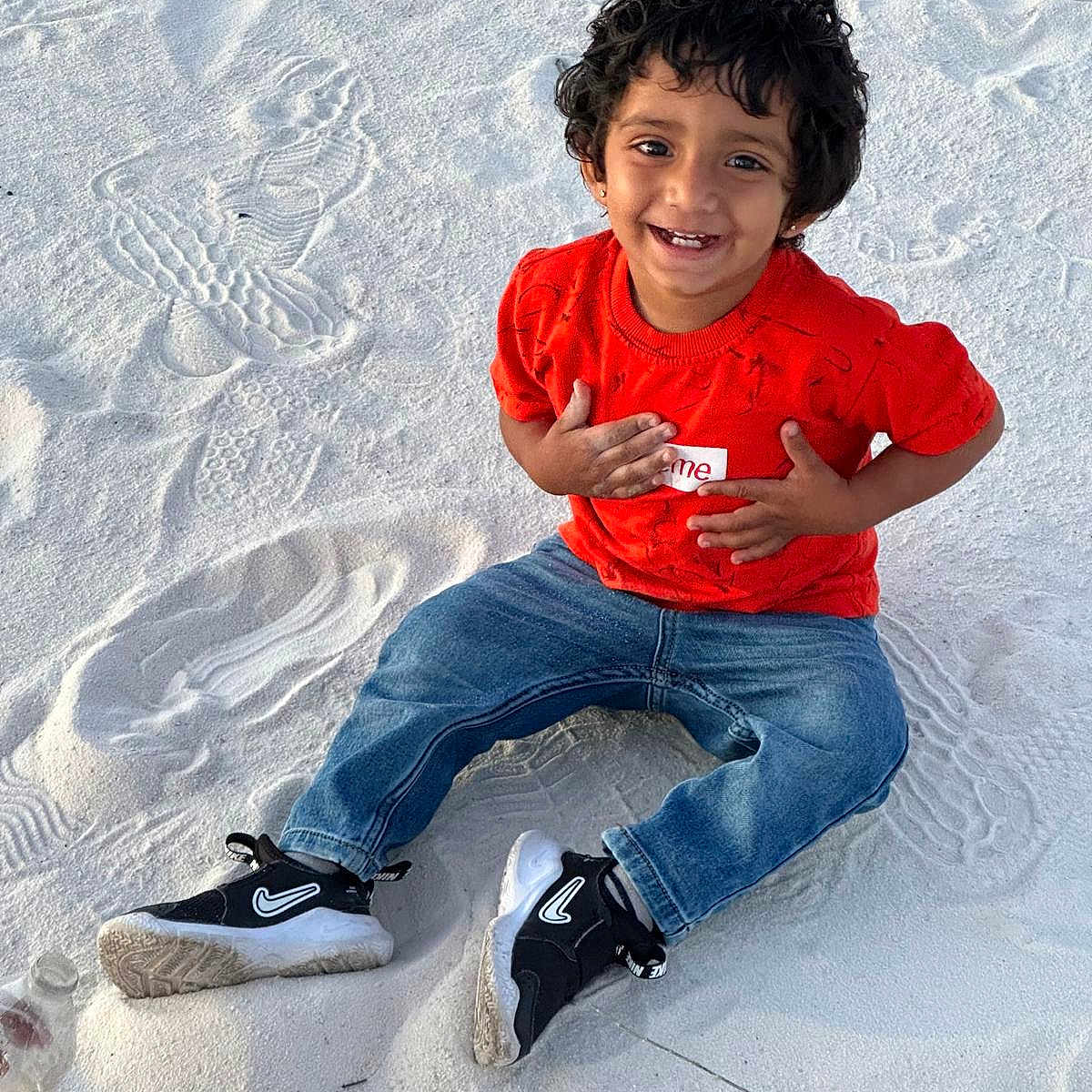 Aadhavan is registered to the contest to win money with this photo: child, smiling, sand, red_shirt, jeans, sneakers, outdoor, happy, curly_hair, casual_clothing, playful, sitting, footwear, nature, daylight, portrait, young_child, joyful, fun, casual