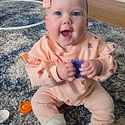 Lily is registered to the contest to win money with this photo: baby, infant, smiling, happy, headband, bow, unicorn_slippers, onesie, peach_clothing, purple_lid, toy_parts, rug, carpet, hardwood_floor, sitting, hands, blue_eyes, indoor, playtime, portrait