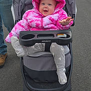 Lily is registered to the contest to win money with this photo: baby, infant, stroller, pink_jacket, hood, tie_dye, smile, happy, outdoor, road, asphalt, gray_stroller, cup_holder, bravo_brand, winter_clothing, sock, adult_leg, walking, parked_cars, hooded_coat