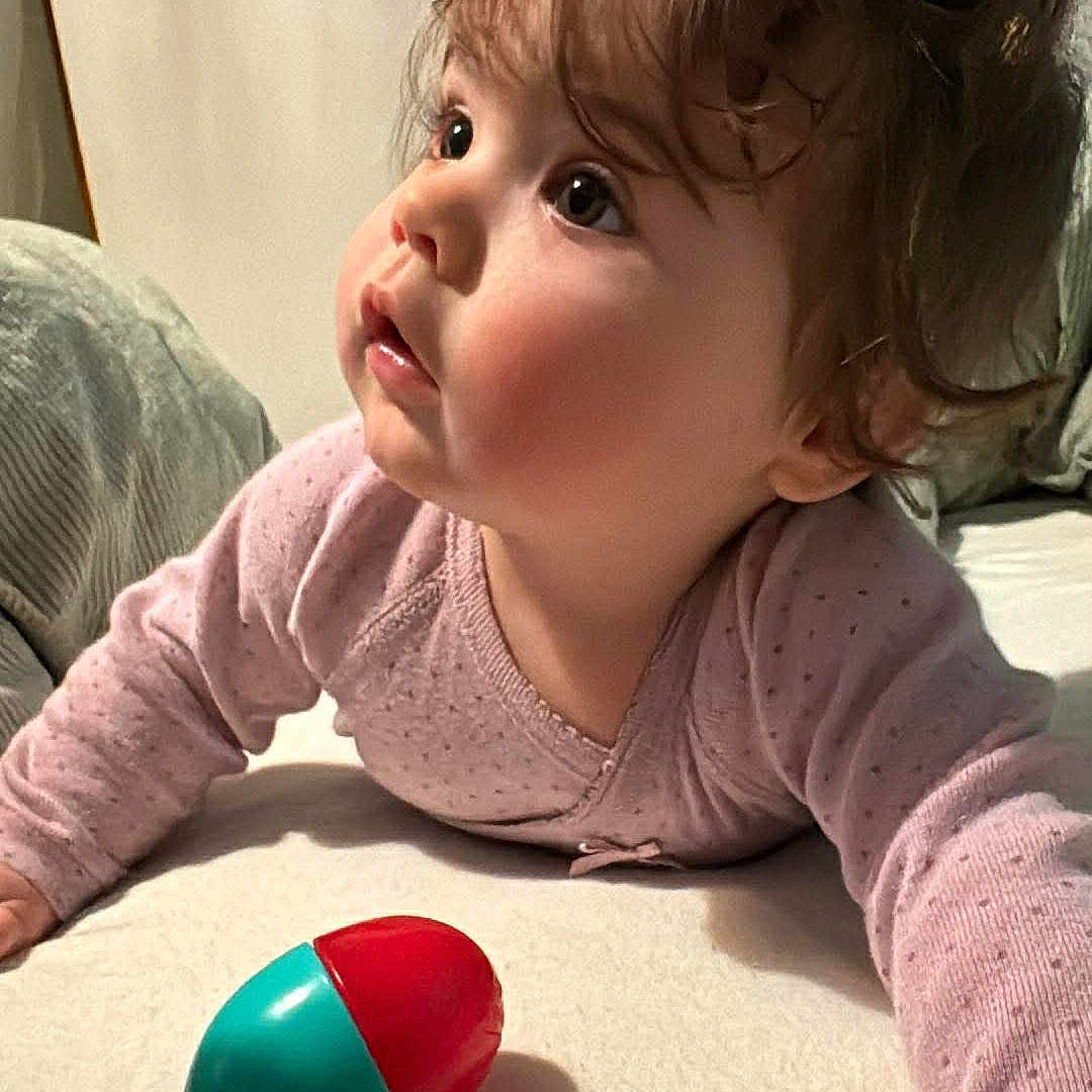 Louisa participe au concours pour gagner de l'argent avec cette photo : baby, bed, bedding, big_eyes, cheeks, closeup, curious_expression, curly_hair, hand, indoor, infant, lying_down, onesie, pillow, pink_clothing, plastic_toy, playtime, portrait, toddler, toy