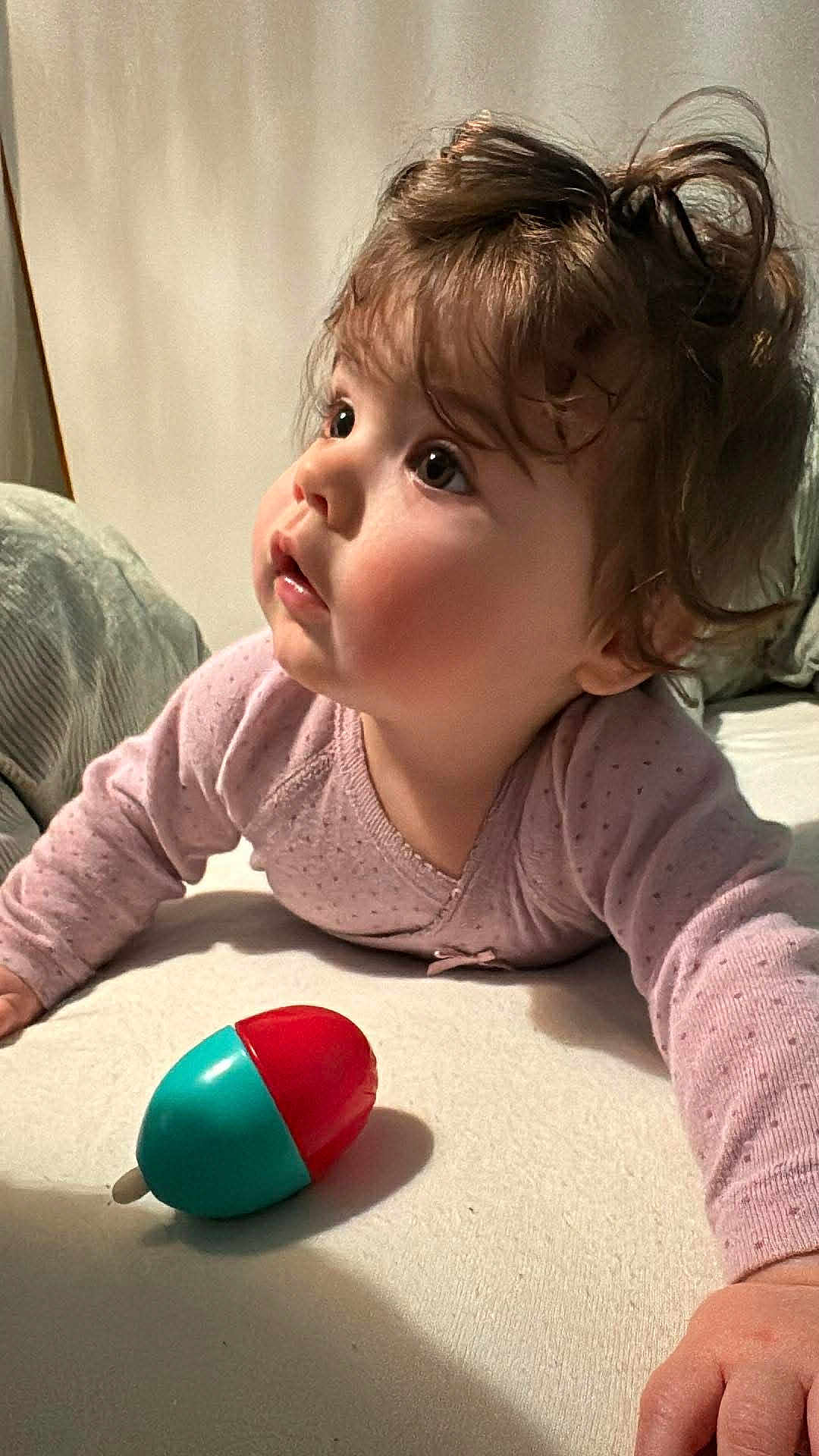 Louisa participe au concours pour gagner de l'argent avec cette photo : baby, infant, toddler, curly_hair, big_eyes, cheeks, onesie, pink_clothing, plastic_toy, toy, bed, bedding, pillow, lying_down, portrait, closeup, indoor, curious_expression, hand, playtime