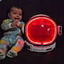 Israel is registered to the contest to win money with this photo: baby, infant, pajamas, colorful_clothing, smiling, face, indoor, couch, dark_background, helmet, astronaut_helmet, red_light, toy, child, sitting, happy, cute, cozy, playful, portrait