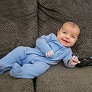 Israel is registered to the contest to win money with this photo: baby, blue_onesie, couch, remote_control, smiling, happy, infant, child, indoor, furniture, cozy, relaxed, person, portrait, seated, cute, home, soft_texture, young_child, content