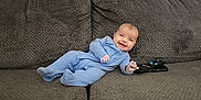 Israel is registered to the contest to win money with this photo: baby, blue_onesie, couch, remote_control, smiling, happy, infant, child, indoor, furniture, cozy, relaxed, person, portrait, seated, cute, home, soft_texture, young_child, content