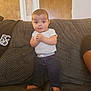 Israel is registered to the contest to win money with this photo: baby, child, standing, couch, furniture, indoors, person, clothing, white_shirt, blue_pants, barefoot, wooden_door, measuring_ruler, home, curious, hands_clasped, expression, young, face, living_room