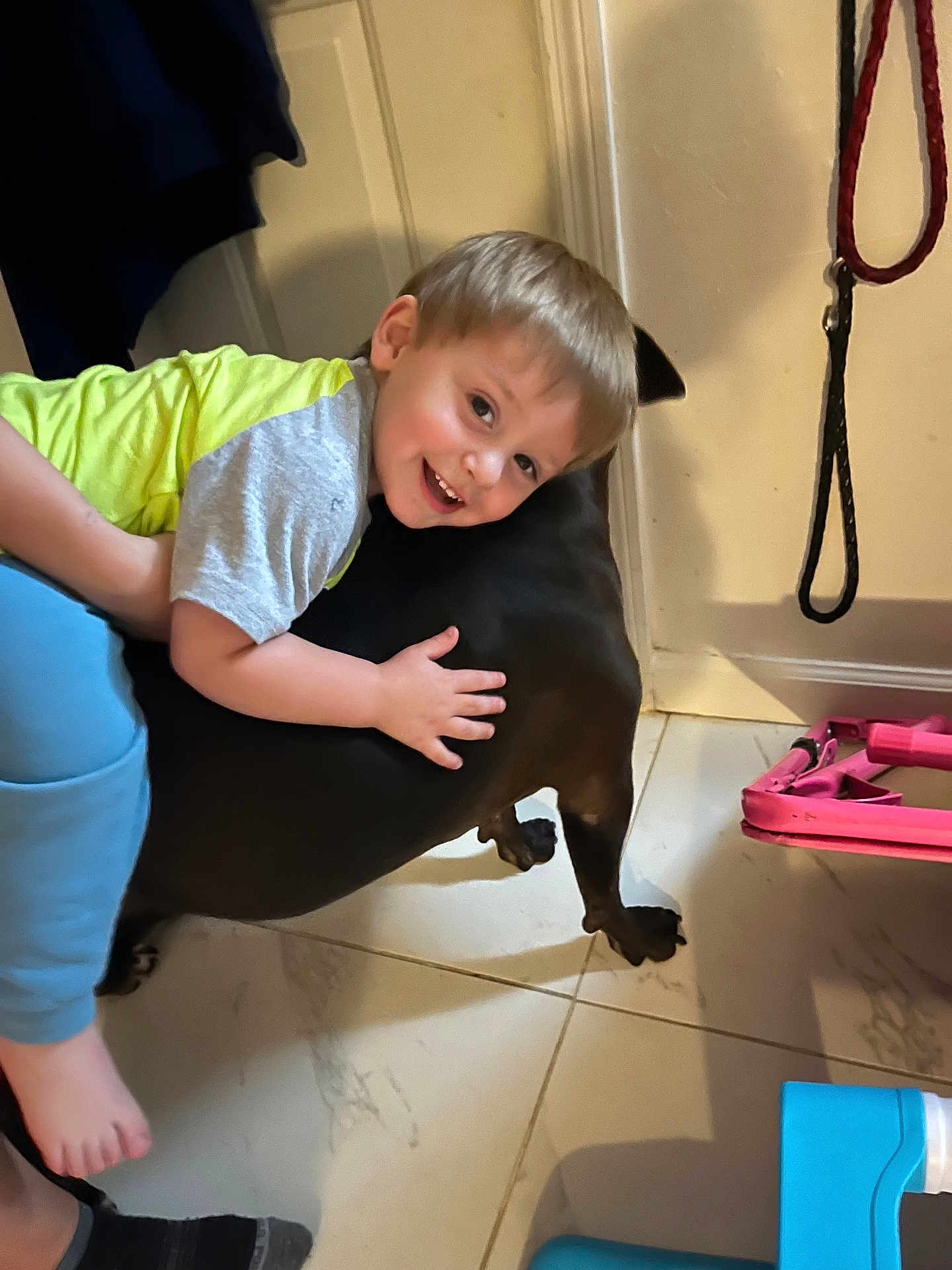 Oliver joined the competition — help win amazing prizes! child, dog, hugging, smiling, indoor, floor, tile, leash, clothing, person, pet, happy, young, light_hair, black_dog, footwear, door, toy, home, flooring