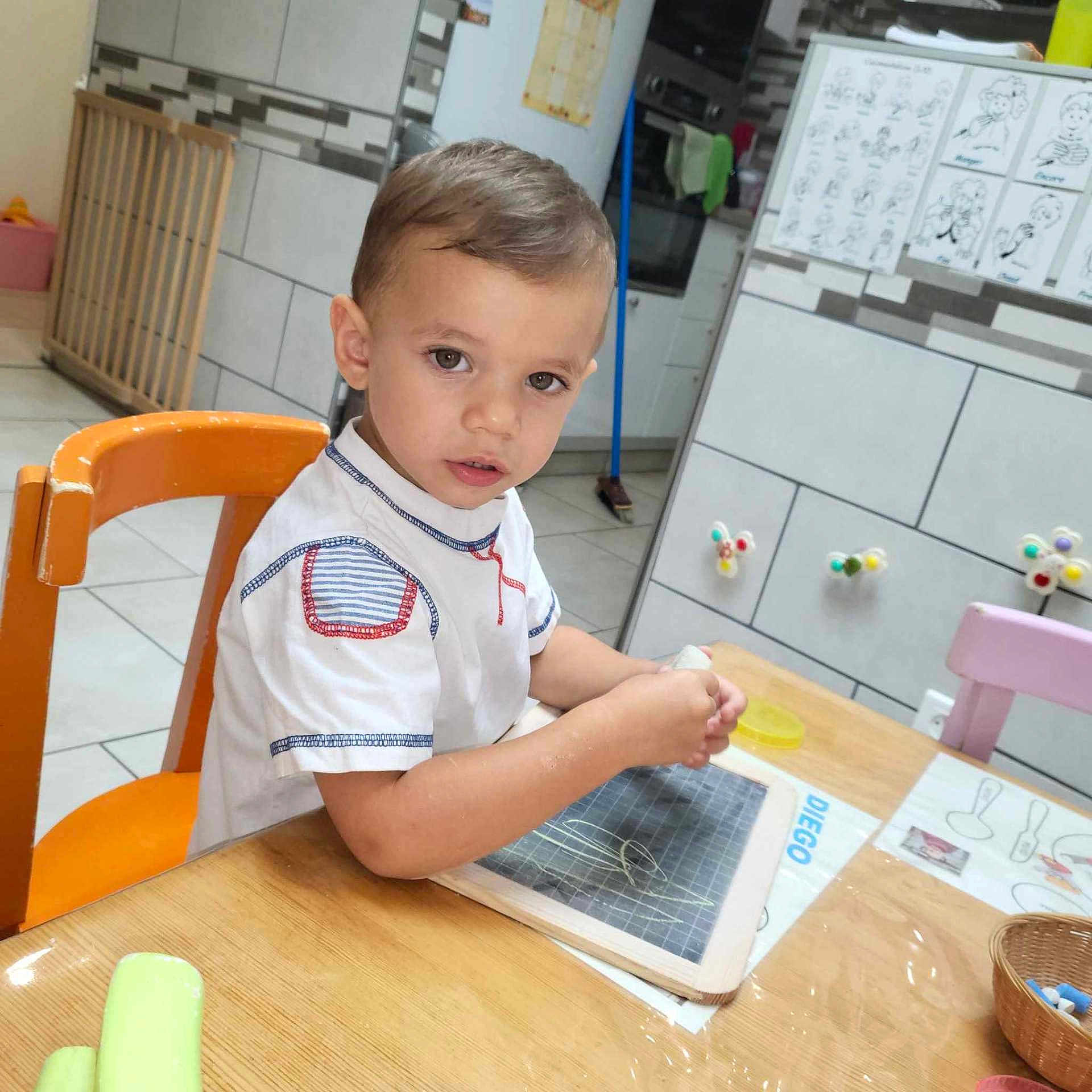 Diego a rejoint le concours — aidez-le/la à gagner de superbes lots ! child, toddler, table, chair, chalkboard, drawing, kitchen, orange_chair, wooden_table, educational_poster, basket, toy, indoor, person, young_child, short_hair, white_shirt, curious, seated, playful