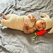 Dior Hendrix is registered to the contest to win money with this photo: baby, infant, knitted_outfit, bunny_ears, carrot_toy, bed, gray_bedspread, lying_down, cute, soft_texture, handmade, portrait, child, cozy, adorable, newborn, face, headwear, indoor, toy