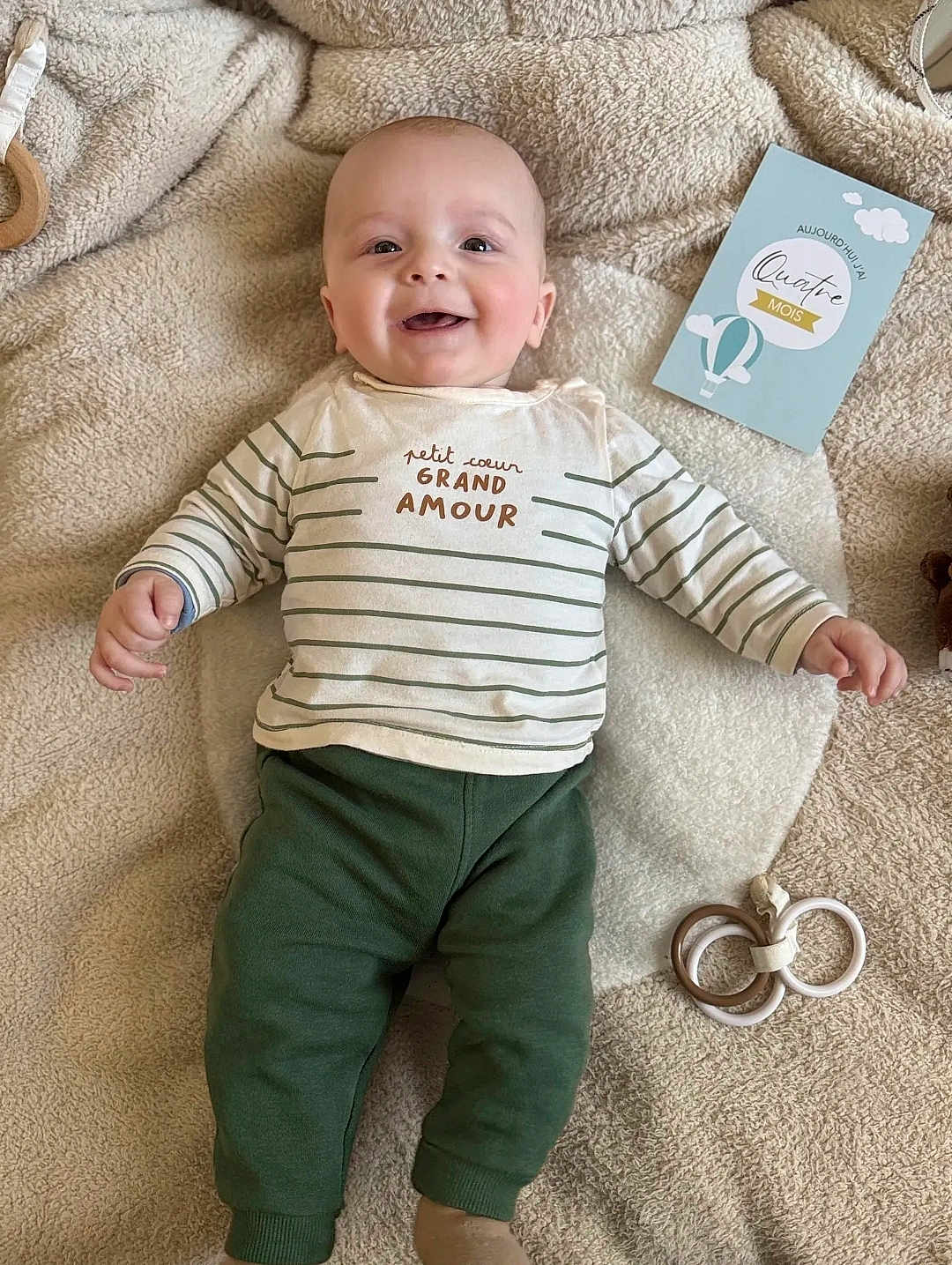 Naël participe au concours pour gagner de l'argent avec cette photo : baby, smiling, infant, striped_shirt, green_pants, blanket, baby_toys, milestone_card, cozy, cute, happy, lying_down, child, face, head, hands, feet, indoors, soft_texture, playful
