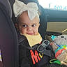 toddler, child, car_seat, headband, bow, toy, car_interior, seatbelt, window, sunlight, clothing, black_outfit, yellow_bib, smile, face, hand, seat, baby, cute, indoors