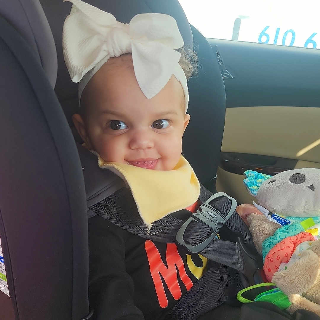 Aziyah joined the competition — help win amazing prizes! toddler, child, car_seat, headband, bow, toy, car_interior, seatbelt, window, sunlight, clothing, black_outfit, yellow_bib, smile, face, hand, seat, baby, cute, indoors