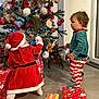 child, christmas_tree, curiosity, decoration, elf_pants, festive, floor, gift_wrap, gingerbread, green, holiday, indoor, lights, ornaments, play, presents, red, santa_costume, toddler, tree skirt