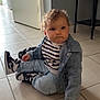 black_shelf, casual_clothing, child, curly_hair, denim_jacket, door, expression, face, floor, home, indoor, jeans, light, person, shelf, sitting, sneakers, striped_shirt, tile_floor, toddler