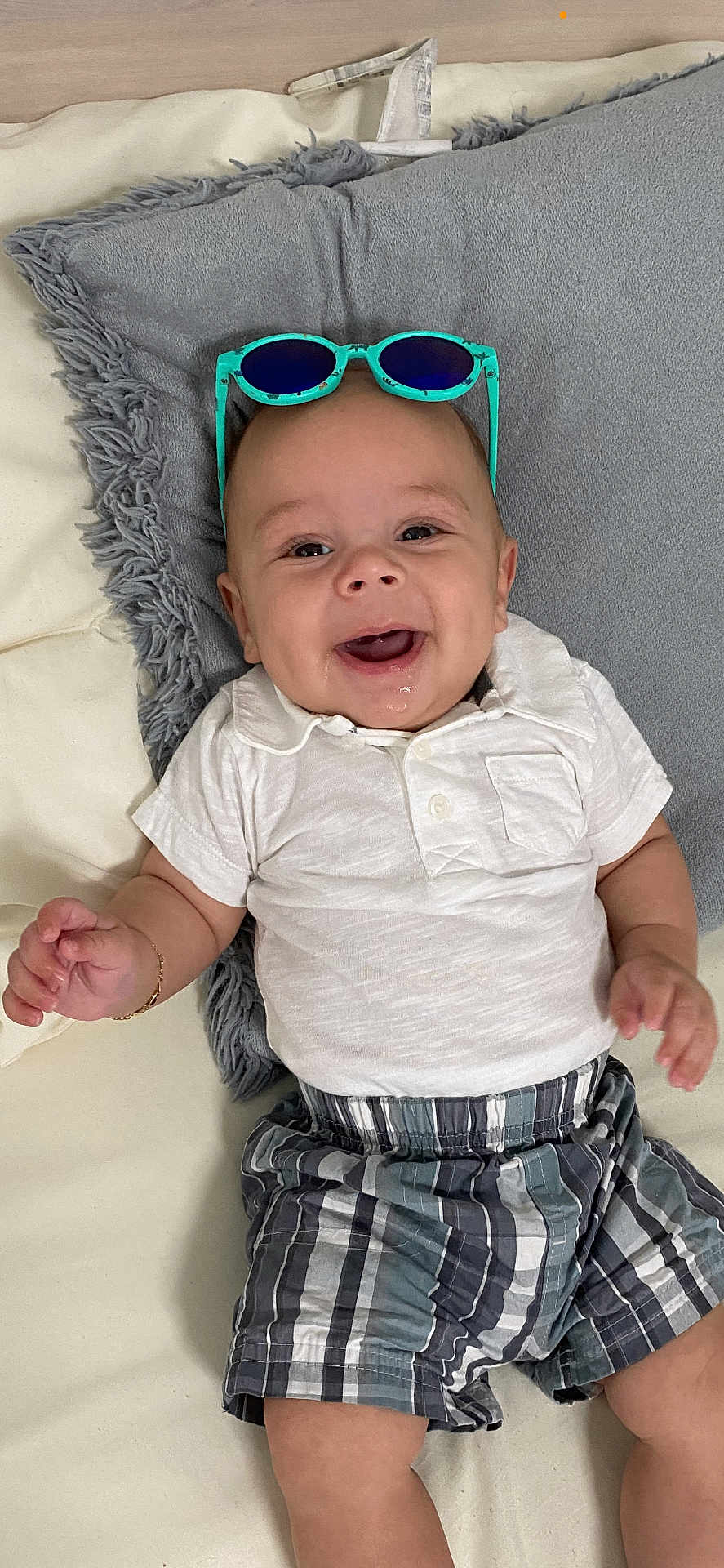 Aaron is registered to the contest to win money with this photo: baby, infant, smiling, sunglasses, plaid_shorts, white_shirt, drool, pillow, bed, happy, child, casual_clothing, lying_down, cute, indoor, person, young, headwear, accessory, toddler