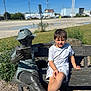 child, boy, statue, bench, outdoor, reading, sculpture, casual_clothing, summer, sunny, blue_sky, grass, plants, urban_background, concrete, casual_pose, daylight, portrait, public_park, curious_expression