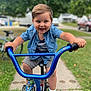 child, toddler, bicycle, blue_handlebars, smiling, outdoor, sidewalk, grass, sneakers, shorts, shirt, happy, play, summer, park, young, person, daytime, recreation, fun
