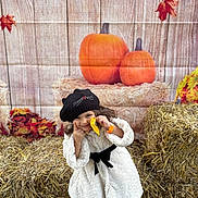 Manisa is registered to the contest to win money with this photo: child, autumn, pumpkin, hay_bale, smile, black_hat, white_coat, fall_leaves, outdoor, seasonal, cute, portrait, festival, snack, playful, orange, decor, person, happy, posing