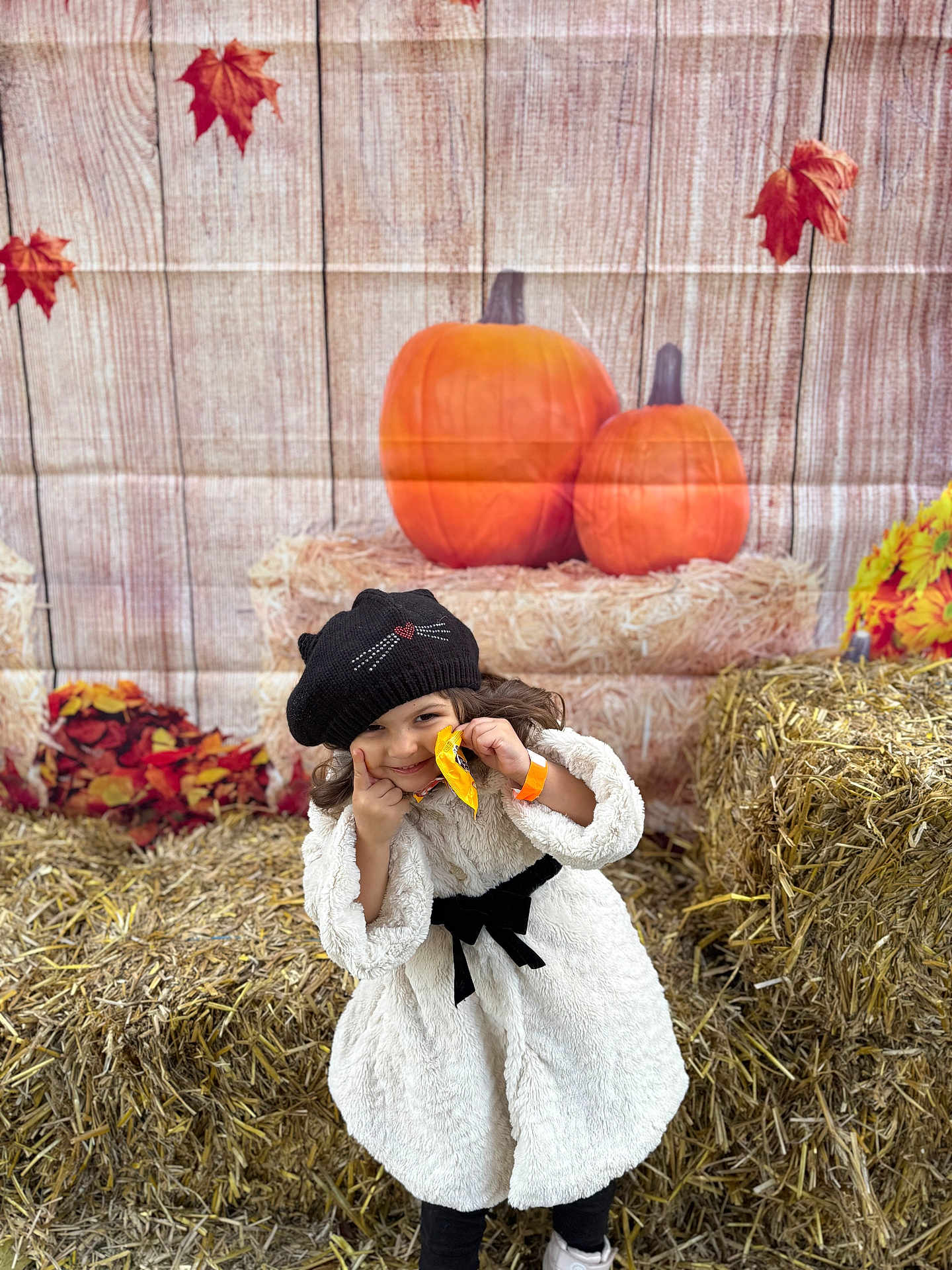 Manisa is registered to the contest to win money with this photo: child, autumn, pumpkin, hay_bale, smile, black_hat, white_coat, fall_leaves, outdoor, seasonal, cute, portrait, festival, snack, playful, orange, decor, person, happy, posing
