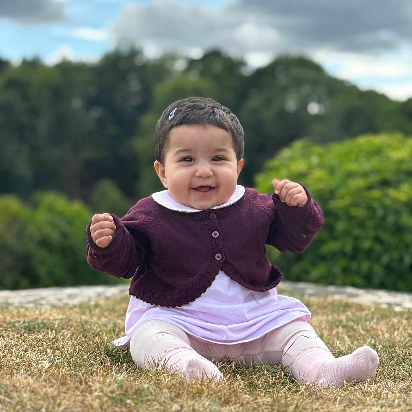 Syra is registered to the contest to win money with this photo: baby, child, clothing, cloudy_sky, cute, face, feet, grass, greenery, hair_clip, happy, nature, outdoor, person, portrait, purple_cardigan, sitting, smiling, white_outfit, young_child