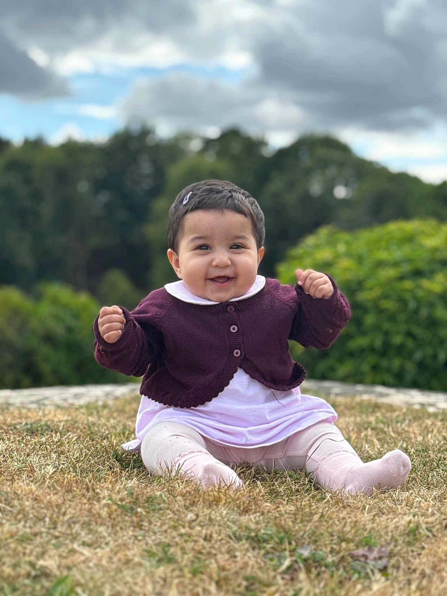 Syra is registered to the contest to win money with this photo: baby, child, smiling, outdoor, grass, purple_cardigan, white_outfit, hair_clip, happy, sitting, nature, greenery, cloudy_sky, cute, portrait, young_child, person, clothing, feet, face