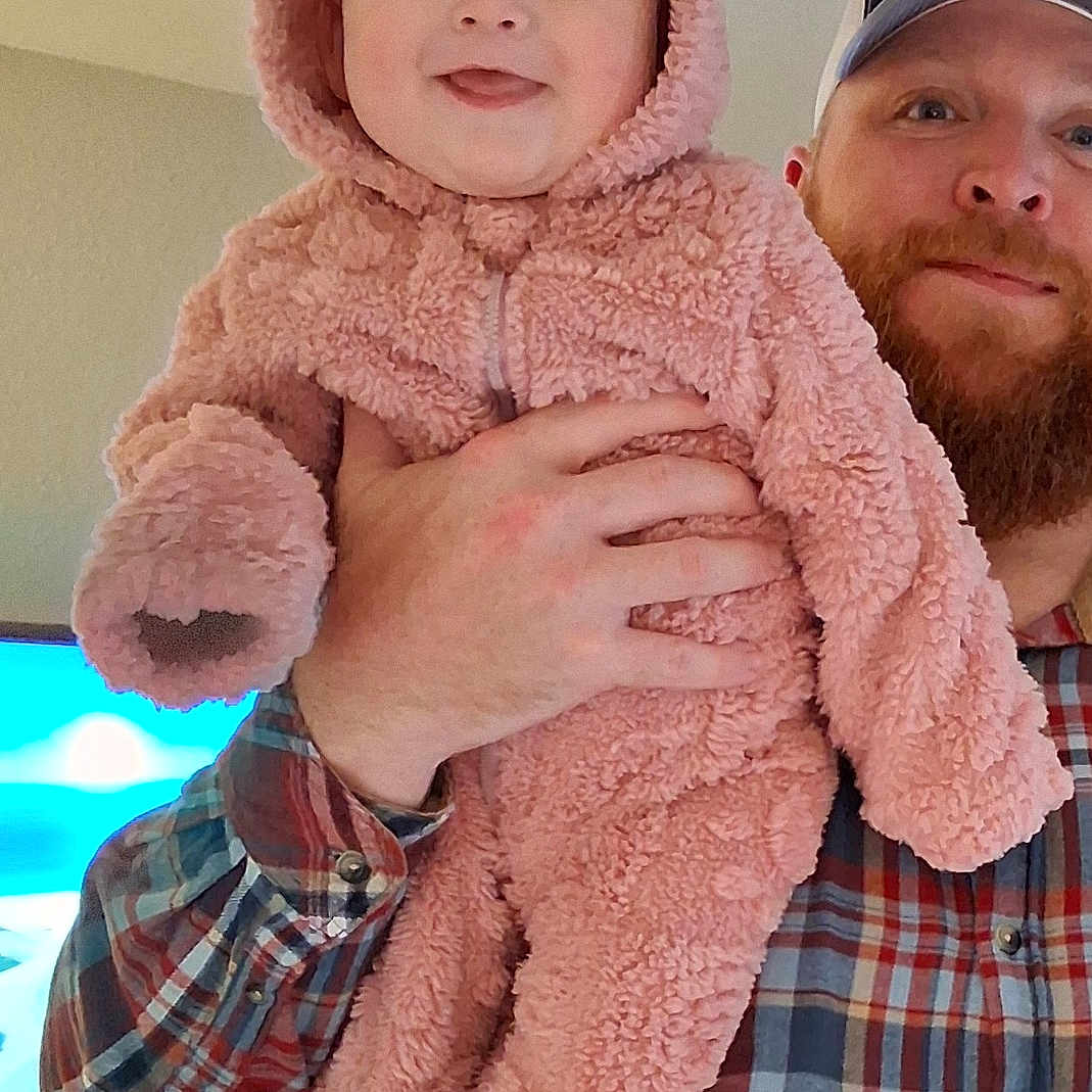 Gabby joined the competition — help win amazing prizes! adult, baby, cap, child, cozy, cute, face, fuzzy_clothing, hand, happy, holding, indoor, living_room, man, person, pink_bear_suit, plaid_shirt, portrait, smiling, television