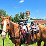 child, toddler, horse, saddle, grass, outdoor, sky, cloud, tree, building, vehicle, fence, sunny, daytime, animal, equine, person, footwear, clothing, nature